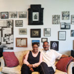 Two people sit on couch in front of wall of photos and documents