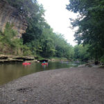 Two kayaks paddle on a river past a large rock wall