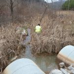 man in green jacket stands in tall grass near small waterway flowing from two large pipes