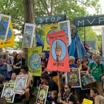 dozens of people hold colorful signs, with a black-and-white "stop mvp" sign above. Part of the capitol building in the background