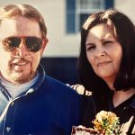 A woman poses next to her husband with her arm around his shoulders.