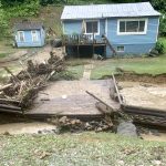 Debris covers one side of a damaged bridge that once connected a blue house to the roadway in the foreground. Receding water reveals badly eroded creek banks.