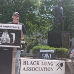 man stands at podium in front of statue of a miner, banner in background says "Black Lung Association"