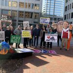 Eighteen women and men line a bricked sidewalk with multistory buildings in the background. They hold signs protesting Gov. Youngkin's attempt to withdraw from the Regional Greenhouse Gas Inistiative.