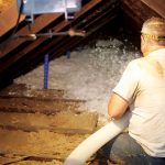 A man in a white shirt with his back to the camera uses a flexible tube to blow insulation into an attic.