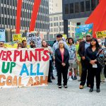 A black woman speaks into a microphone. People stand behind her holding signs calling for coal ash cleanup. A large banner held by several people at the front of the group reads, "Make Power Plants Clean Up All Their Coal Ash."