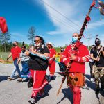 A crowd of Indigenous people, some dressed in traditional clothing or carrying banners, walk along a road behind a woman carrying a vessel of water.