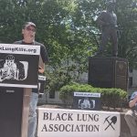 A man in a black t-shirt speaks at a podium behind a sign depicting a skeleton wearing a miner's hat that says BlackLungKills.org in black lettering. A banner that says Black Lung Association is draped over a table behind him and a statue of a miner rises above them in the background.