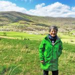 A woman with short, graying hair wearing pants and a long, green jacket stands in a pasture with fields and rolling hills behind her.