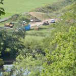 Heavy equipment and vehicles are seen in a fenced off area of disturbed land next to a river.