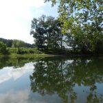 Drilling equipment can just barely be seen in acleared area behind a placid river reflecting clouds and trees.