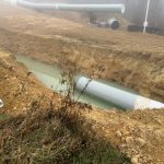 A large, partially buried, green pipe lays half submerged in water in a trench. Other pipe is visible off to the sides of the trench.