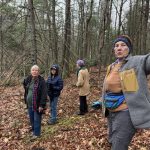 Four women of various ages are standing on a slope in a forested area. Trees are bare except for evergreens and the ground is littered with fall foliage in browns and dull reds. The women are wearing cool weather clothes and one looks like she is speaking toward the camera while leaning one arm against a tree trunk.