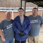 A young man in a blue jacket stands between two older men wearing gray t-shirts. the roof of the brick building behind them is covered in solar panels.