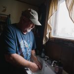 A white-bearded man in a blue t-shirt stands by a sink with water running from the faucet.